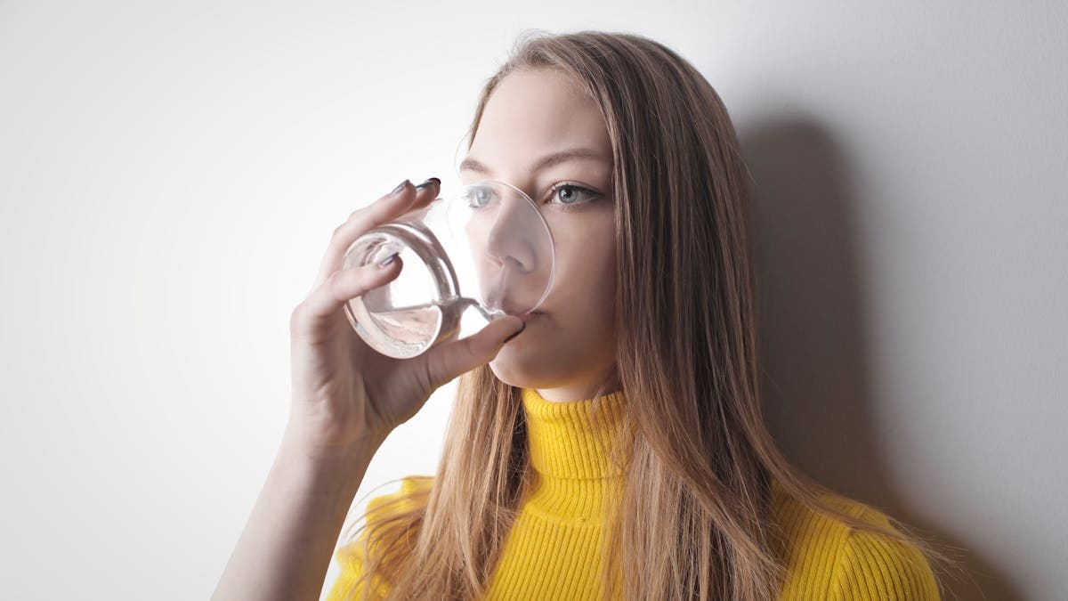 Young woman in a yellow top, drinking water from a glass against a white wall.