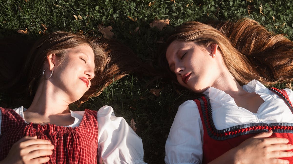 Twin sisters lie on grass wearing traditional dirndl dresses in Graz, Austria.